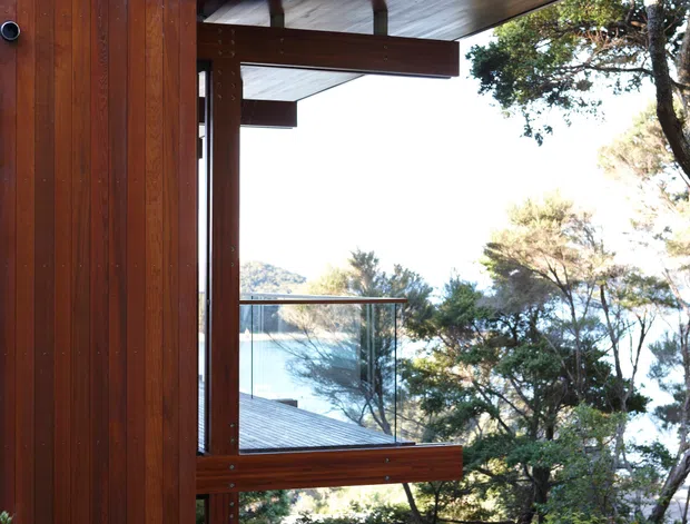 Exterior architectural detail of a contemporary timber-clad house set among dense garden vegetation, with a projecting flat roof or canopy, exposed timber framing, and large glazed openings. A glass balustrade overlooks a wooded, possibly waterfront landscape visible beyond the structure.