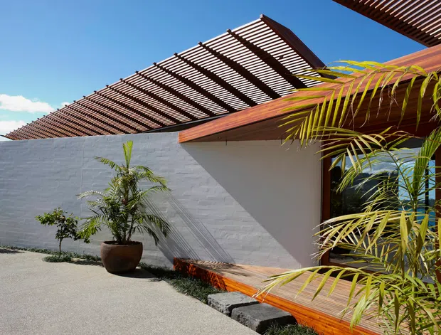 Exterior view of a low-profile modern building with white rendered masonry walls, warm timber decking and projecting timber pergola screens. Tropical palms and sparse landscaping frame the shaded entry or terrace, with strong sunlight and crisp geometric shadows.