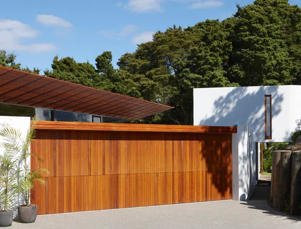 Exterior view of a modern residential property, featuring a broad warm timber garage or privacy door, a low sweeping terracotta tiled roof, and a crisp white cubic upper volume with a narrow vertical window. The house is set among mature trees and a landscaped, slightly sloping site, with a paved driveway in the foreground.