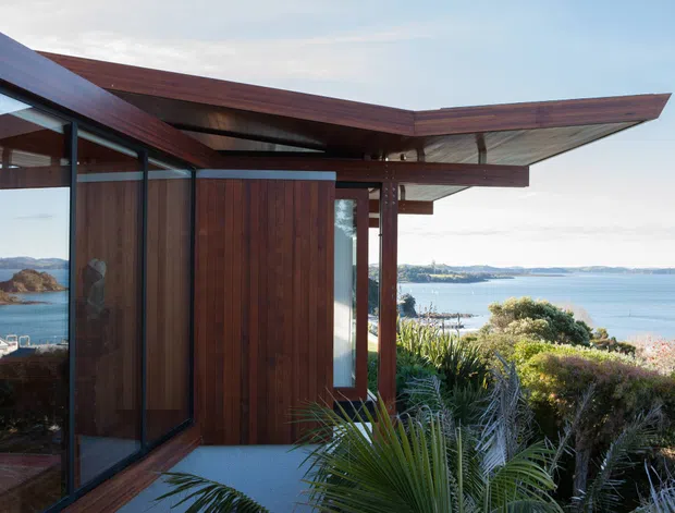 Exterior view of a modern coastal residence with dark timber-clad walls, expansive floor-to-ceiling glazing, and dramatic cantilevered roof planes. The house is set above a bay or waterfront, with native coastal planting in the foreground and distant shoreline visible beyond.