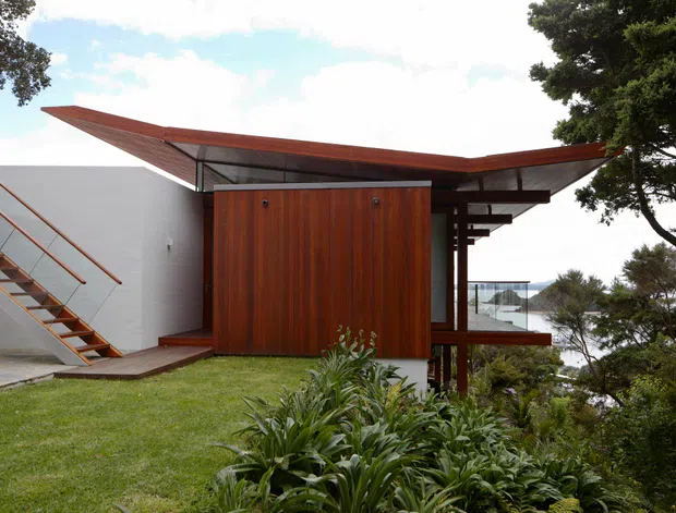 Exterior view of a modern coastal house with a dramatic low-pitched, wing-like roof, warm vertical timber-clad central volume, white rendered walls, exposed timber supports, and an external open-riser stair. The house is set within landscaped planting overlooking a visible body of water.