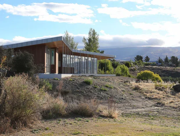 A low-profile contemporary building set in a sparse, rugged landscape. The structure combines a broad flat or shallow-sloping roof, dark vertical cladding, warm timber-toned wall sections, and an extended glazed facade with slender vertical frames. Dry grasses, scrub vegetation, open ground, and distant mountains create a strong rural or alpine setting.