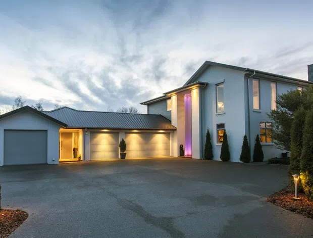 Exterior photograph of a contemporary two-storey residence at dusk, with a light rendered facade, dark metal low-pitched roofs, attached single-storey garage, broad paved driveway, tall narrow windows, and a dramatic illuminated vertical entry feature. Landscaping includes clipped hedges and conical evergreens.