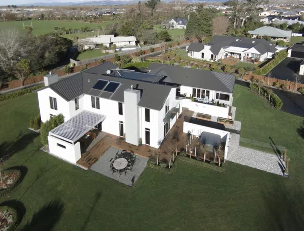 Aerial view of a large contemporary white multi-level residence with dark pitched roofs, extensive glazing, solar panels, terraces, a pergola-covered outdoor area, paved paths, and expansive landscaped lawns. The home sits in a suburban/rural-edge neighbourhood with surrounding houses and open fields.