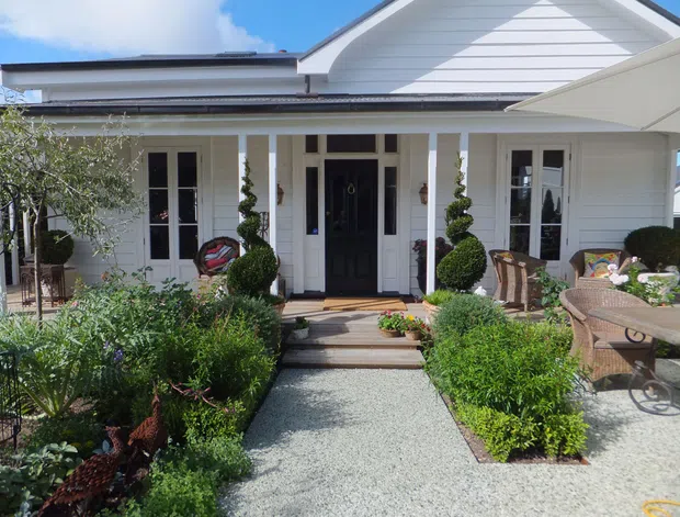 Bright front exterior of a single-storey home with a white horizontal weatherboard facade, gabled roof, covered entry porch, dark glazed front door, and symmetrical tall windows. A gravel path leads through a neatly landscaped garden with clipped shrubs, topiary, and outdoor wicker seating.