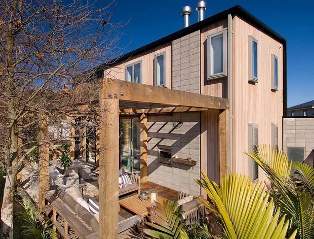 Exterior view of a compact multi-storey contemporary home with a warm timber-clad upper volume, pale masonry or concrete-block wall panels, dark roof edging, and tall narrow windows. A timber pergola frames a decked outdoor living area with seating and tropical planting in the foreground.