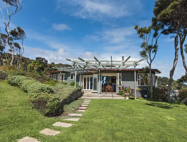 Exterior view of a low-rise contemporary coastal-style house set within a landscaped garden, with extensive glazing, a pergola-like exposed roof structure, and a stepping-stone path leading to a covered patio entrance. Wind-shaped trees and open lawn surround the dwelling.