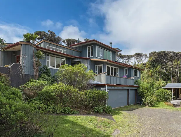 Exterior view of a multi-level residential house set within dense coastal-style landscaping. The home has dark vertical cladding, contrasting light window trim, broad glazed openings, projecting roof forms, and an attached lower garage. A small separate pavilion or shed sits to the right, with a gravel driveway and planted garden in the foreground.