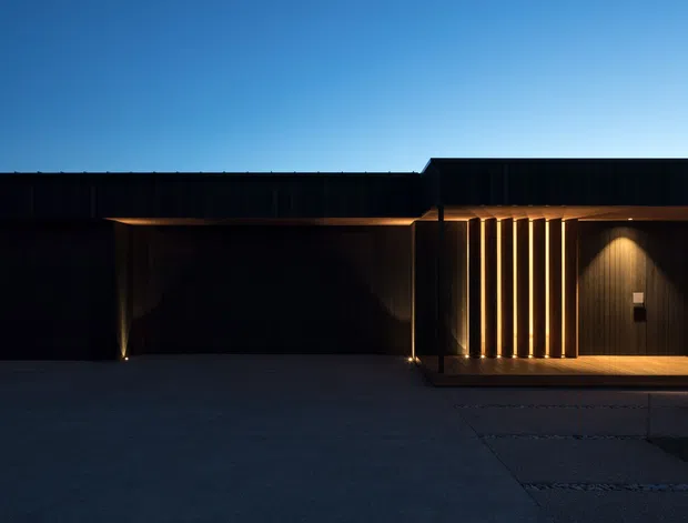 A dark, minimalist contemporary building facade photographed at dusk. The low, elongated structure features black vertical cladding, a flat roof, concealed warm architectural lighting, and a recessed entry or covered porch screened by evenly spaced vertical timber or metal battens. A broad paved forecourt occupies the foreground, with a deep blue evening sky above.