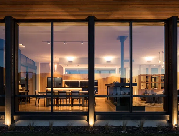 Dusk exterior view through full-height glazing of a contemporary home, revealing a warm open-plan interior with a dining table, kitchen cabinetry and island, living area, and dark structural window framing. The exterior is clad or finished in dark tones with timber-lined soffit or ceiling above the glazing and subtle landscape lighting along the base.