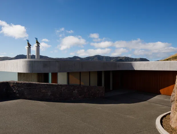 Low-profile modern residence integrated into a sloping coastal site, viewed from the driveway. The building has a broad flat roof with a projecting concrete fascia, dark timber-clad walls, extensive recessed glazing, twin metal flues, and a curved dark stone retaining wall. Hilly terrain and water are visible beyond the house.