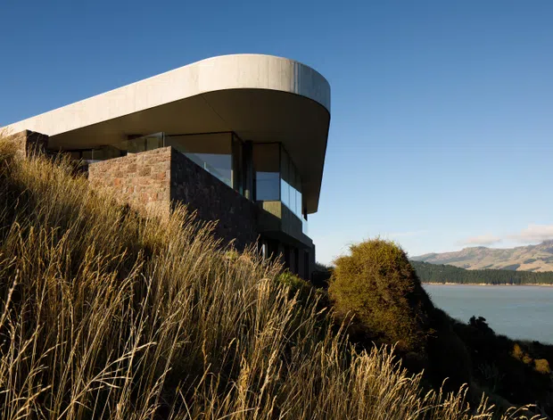 Exterior architectural photograph of a contemporary house set into a sloping, grassy lakeside landscape. The building features a sweeping curved flat roof with a pale smooth finish, dark-framed glazing, and substantial irregular stone walling. A lake and mountainous terrain are visible beyond the property.