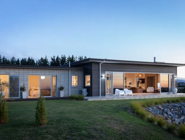 Single-storey contemporary residential home viewed from the landscaped exterior at dusk. The long, low building has dark horizontal cladding, extensive floor-to-ceiling glazing and sliding doors, a shallow overhanging roof, and a partially enclosed covered terrace with outdoor seating. A lawn, planted trees, rocky drainage or retaining edge, and distant hills surround the house.