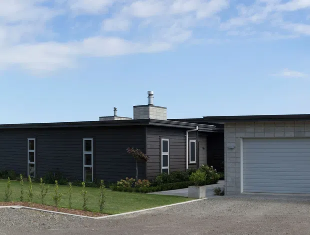 Single-storey contemporary residential house with a long dark horizontal facade, narrow vertical windows, low-pitched roof, and an attached light-grey rendered or masonry garage volume with a wide sectional garage door. The home sits beside a neatly landscaped lawn and gravel driveway under a bright sky.