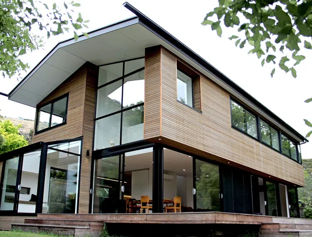 Two-storey contemporary residence viewed from the garden, with a sharply pitched asymmetrical roof, extensive floor-to-ceiling glazing, black-framed windows and doors, and horizontal timber-clad upper walls. The open lower level reveals a dining area, while surrounding trees and a sloping landscape frame the house.