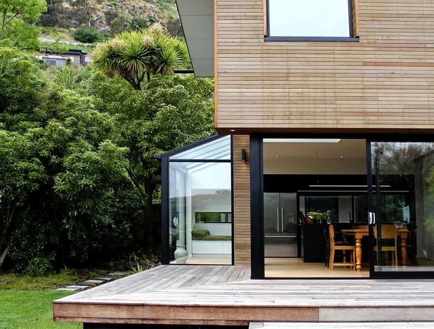 Exterior view of a contemporary two-storey house set against a landscaped, sloping hillside. The facade combines horizontal timber cladding with dark-framed glazing, including large sliding doors opening to a raised timber deck. A glazed corner extension or sunroom is visible beside the main volume, with a dining area seen inside. The deck has broad steps descending to a lawn and garden.