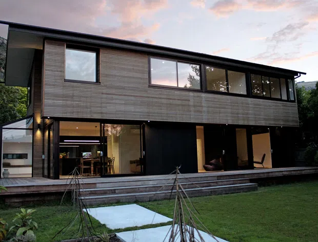 Exterior view of a contemporary two-storey home set within a landscaped garden. The building has a strong horizontal form, dark low-pitched roof, extensive black-framed glazing, and an upper level clad in narrow horizontal timber boards. A covered ground-floor terrace and open-plan living spaces are visible through the glazing, with a paved path leading across the lawn.