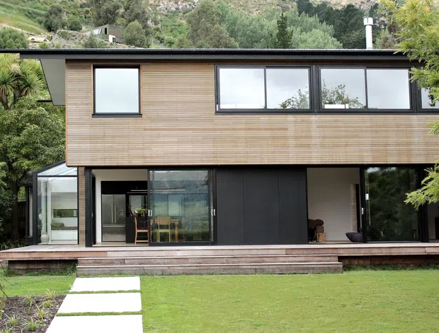 Exterior view of a contemporary two-storey house set into a lush, wooded hillside. The upper level is clad in horizontal light timber boards with a thin flat roof and a row of dark-framed windows, while the lower level combines dark cladding, extensive glazing, and open living areas connected to a timber deck. A glass-enclosed extension is visible on the left, with a lawn and stepping-stone path in the foreground.