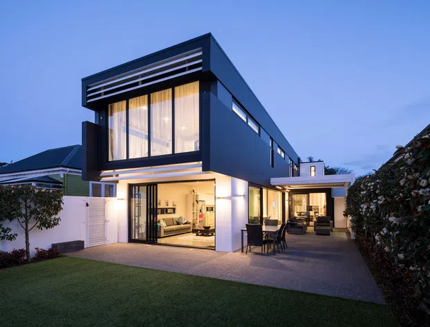 Twilight exterior view of a contemporary two-storey home with a dark, box-like upper volume, extensive glazing, horizontal louvres, and warm interior lighting. A covered outdoor dining and lounge area extends beside the house, with a paved forecourt, lawn, hedging, and neighbouring homes visible around it.
