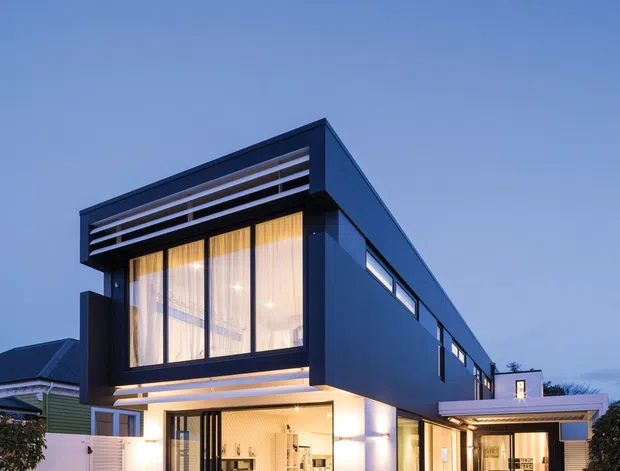 Twilight exterior view of a contemporary multi-level residence with a bold box-shaped upper volume, dark blue-grey cladding, extensive glazing, horizontal louvres, and illuminated ground-floor living spaces. A covered outdoor entertaining area with dining furniture and lounge seating extends from the house, opening onto a paved terrace and lawn.