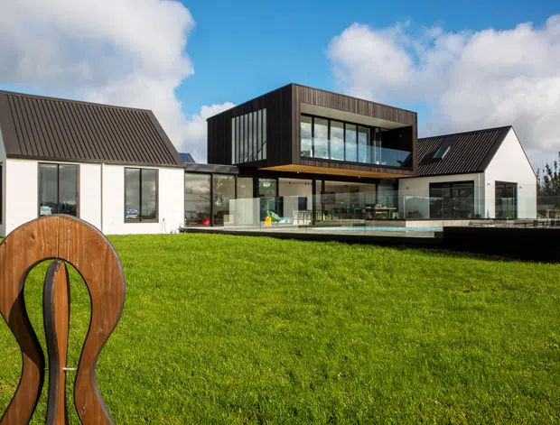 Exterior view of a contemporary multi-wing residence set behind a broad lawn. The house combines white rendered gabled volumes with dark metal pitched roofs and a prominent dark-clad upper-level box featuring extensive floor-to-ceiling glazing. A glazed ground-floor living area opens toward a dark deck or terrace, while a large abstract timber sculpture stands in the foreground.