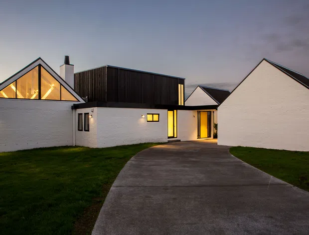 Exterior view of a contemporary single-storey residential complex at dusk. White masonry walls, sharply pitched gable forms, dark vertical timber-clad upper volumes, and warm interior lighting create a strong contrast. A curved concrete driveway leads toward the recessed entrance, with lawn on either side.
