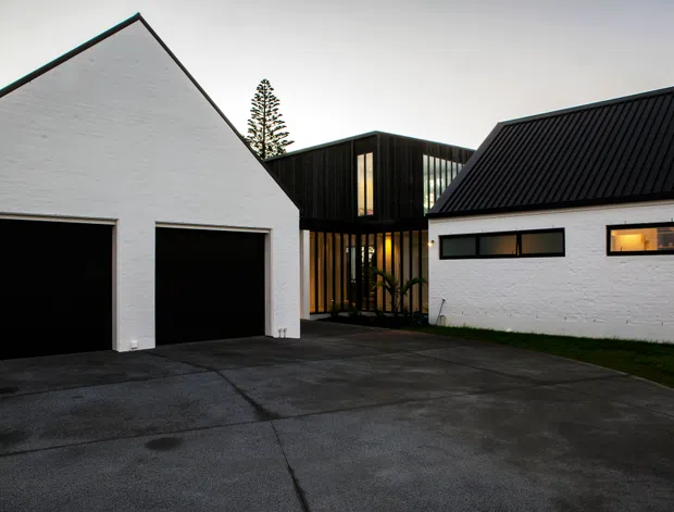 Exterior view of a contemporary residential compound with white-painted brick gabled garage buildings, black ribbed metal pitched roofs, and a dark timber-clad glazed connector at the centre. The broad concrete driveway dominates the foreground, with warm interior lighting visible through narrow windows and glazing.