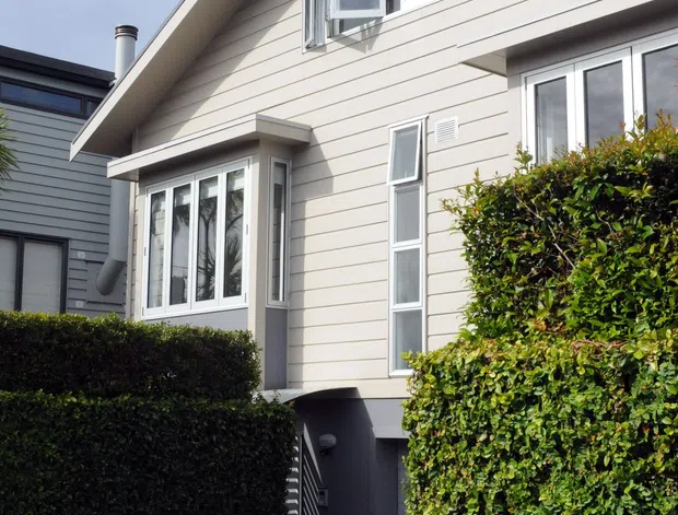 Exterior view of a compact multi-level suburban house with pale horizontal weatherboard cladding, a steep gabled roof, white-framed casement windows, projecting bay-style glazing, and neatly trimmed hedge screening. A lower-level garage or entry area is partially visible beneath the main living level.