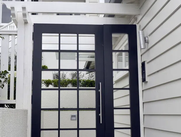 A close-up exterior view of a residential entry courtyard featuring a dark navy glazed entry door with multiple rectangular panes and a matching narrow sidelight. White horizontal weatherboard cladding, white pergola beams, porch framing, and a gabled upper facade create a crisp, light-toned entrance. The image also shows a concrete-paved approach and glimpses of planted courtyard areas through the glazing.
