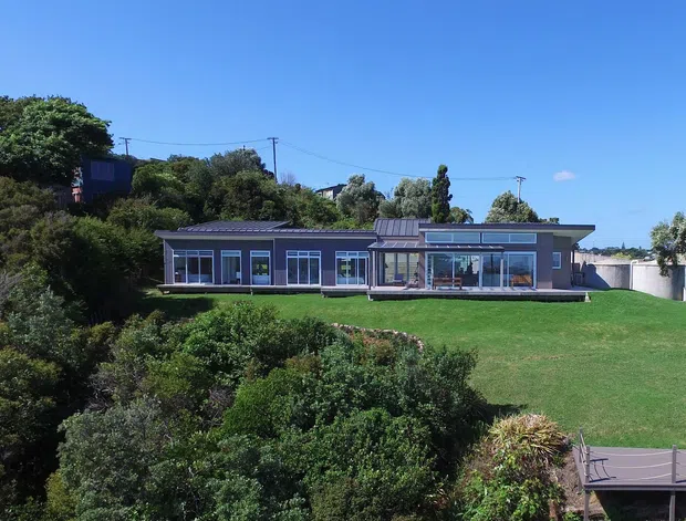 Single-storey contemporary house set on a landscaped sloping site, with a long low-profile form, dark metal roof, extensive floor-to-ceiling glazing, and a broad lawn overlooking dense planting. The image is an exterior architectural view in clear daylight.