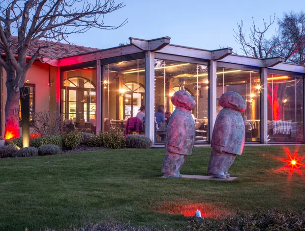 Twilight exterior view of a low-rise contemporary building with extensive floor-to-ceiling glazing and a covered outdoor dining or lounge area. The landscaped lawn features two large sculptural figures and dramatic red accent lighting, while warm interior lighting is visible through the glass.