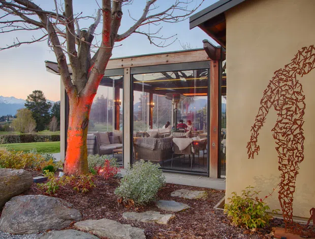 Exterior view of a contemporary glazed dining or living pavilion attached to a plastered building, set within a landscaped garden. Large floor-to-ceiling glass panels reveal wicker seating, a dining table, pendant lighting and warm interior illumination. Natural stone stepping slabs, boulders, planting beds and a mature leafless tree frame the facade, with distant mountains visible beyond the property. A decorative metal wall artwork is mounted on the tan exterior wall.
