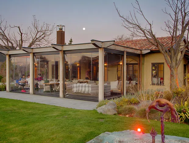 Exterior view of a single-storey home at dusk, featuring a glazed sunroom or enclosed outdoor living extension with slender structural framing, a flat roof, and open views into a furnished living area. The adjoining house has warm-toned rendered walls and a terracotta tiled pitched roof, set within a landscaped garden.