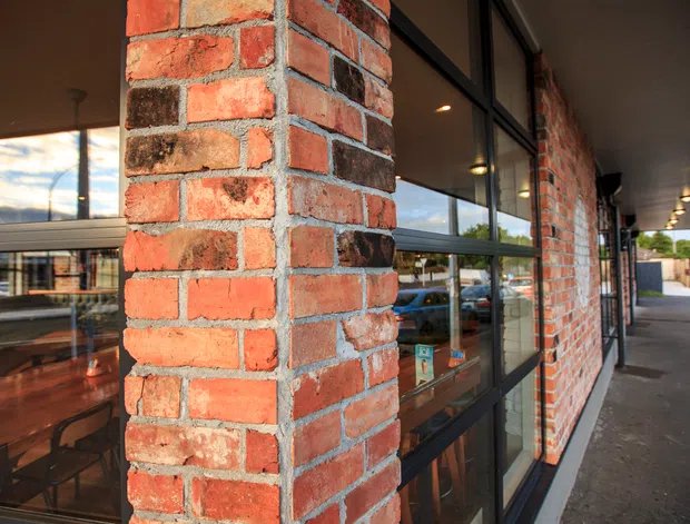 Exterior view of a commercial-style building with red and weathered brick columns, dark-framed floor-to-ceiling glazing, and a covered pedestrian arcade. Interior dining or retail furnishings are visible through the windows and reflections.