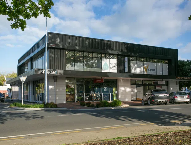 Street-facing two-storey commercial building with a dark corrugated upper facade, extensive glazed shopfronts, and ground-floor retail tenancies. The building fronts a paved urban road with parked cars and mature street trees.