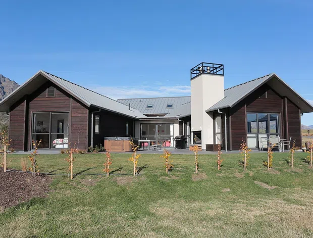 Exterior view of a contemporary single-storey residence with two dark timber-clad gabled wings connected by a central low-pitched section. The home features grey metal roofing, extensive glazing, a prominent white chimney with a black metal cap, and landscaped lawn with young orchard plantings. Mountainous terrain is visible in the background.