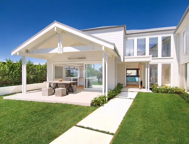Bright exterior view of a white, multi-wing residential home with horizontal weatherboard cladding, gabled roof forms, large glazed doors and windows, and a covered outdoor dining or entertaining pavilion opening onto a manicured lawn. A concrete path leads through the landscaped courtyard toward the house.