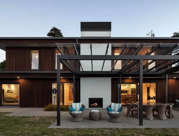 Exterior view of a contemporary two-storey home with dark vertical timber cladding, broad flat rooflines, extensive glazing, and a steel-framed covered outdoor entertaining area. The covered patio includes lounge seating, a central outdoor fireplace, and a dining setting, while illuminated interior spaces are visible through the glazing.
