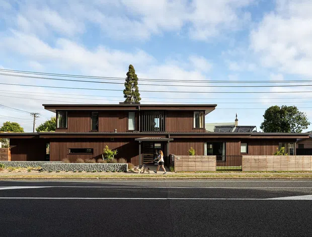 Street-facing view of a contemporary two-storey residential house with a long, low horizontal form. The facade combines dark timber-look vertical cladding, large glazed openings, projecting flat roof planes, and a timber slat fence. A gravel landscape strip and low stone retaining edge sit between the home and the broad paved road, with neighbouring houses and overhead utility wires in the background.