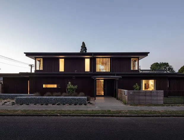 Street-facing exterior photograph of a contemporary two-storey residence at dusk. The house has a strong horizontal composition, a flat projecting roof, dark vertical timber-like cladding, warm illuminated windows, a recessed central entry, and low front landscaping with timber fencing and masonry edging.