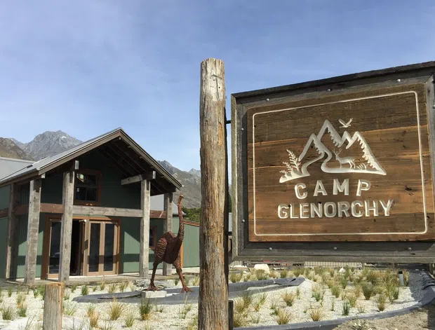 Exterior view of a rustic alpine camp entrance, featuring a timber lodge-style building with a steep gabled roof, covered porch, dark green and natural wood finishes, and a large weathered wooden sign reading “Camp Glenorchy.” Mountain peaks are visible in the background, with landscaped gravel and native-style planting in the foreground.