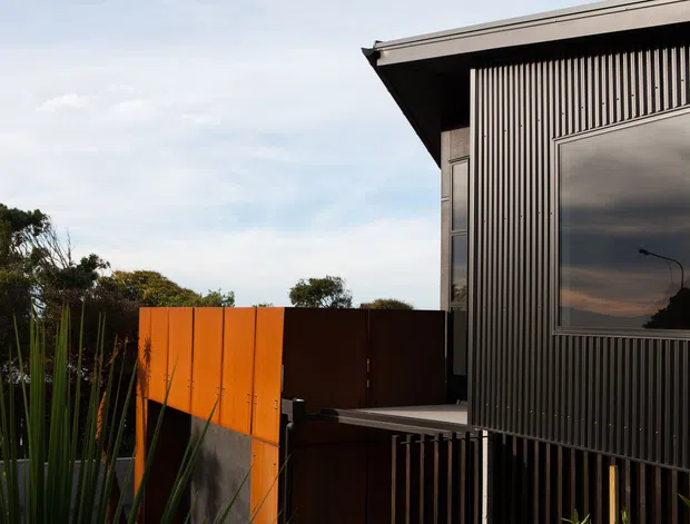 Exterior view of a contemporary dark-clad building with a sharply projecting flat roof edge, vertical ribbed facade panels, and a large reflective window. A vivid orange privacy or boundary screen contrasts with the charcoal exterior, with landscaped planting visible along the side.