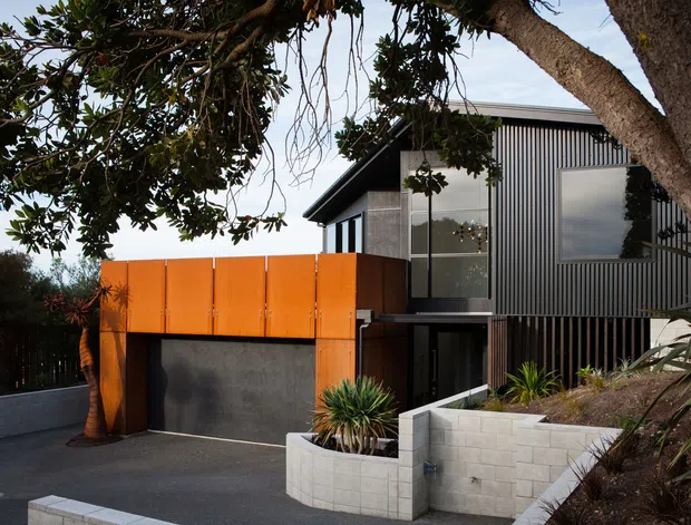 Exterior view of a contemporary multi-level residence set into a sloping, landscaped site. The house features dark vertical ribbed cladding, large glazed openings, a vivid orange projecting garage volume with a dark garage door, and pale rendered or concrete-block retaining walls. Mature trees and planted beds frame the driveway and entrance.