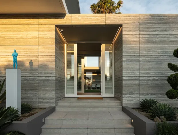 Front entrance of a refined modern residence, featuring a broad horizontally banded stone or concrete-clad facade, deep recessed entry, glazed double doors with louvred side panels, broad steps, and sculptural drought-tolerant landscaping with a pedestal sculpture.