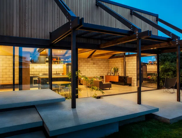 Exterior dusk view of a contemporary multi-level residence with a dramatic exposed dark steel post-and-beam structure, vertical timber cladding, broad glazing, and projecting concrete terraces. Warmly lit interiors are visible through the openings, including a kitchen/dining area and a living room, while the covered outdoor terrace extends across the foreground.