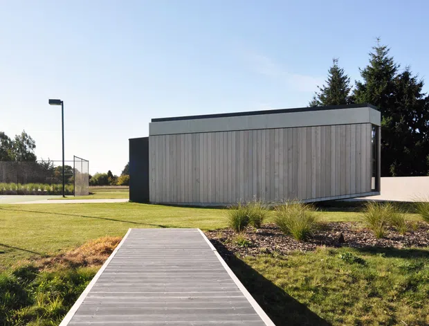 A minimalist single-storey pavilion-like building in a landscaped outdoor setting. The rectangular structure has a flat roof, dark side walls, and a prominent facade clad in vertical pale timber boards. A timber boardwalk leads toward the building through lawn and ornamental grasses, with a sports court or recreational area visible in the background.