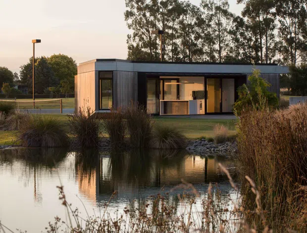 A compact contemporary flat-roof pavilion set beside a landscaped pond. The building has dark vertical exterior cladding, large black-framed glazed openings, and a warm illuminated interior visible through the glazing. Native grasses, rocks, and reflections in the water form the foreground, while tall trees and simple pathway lighting sit behind the structure.