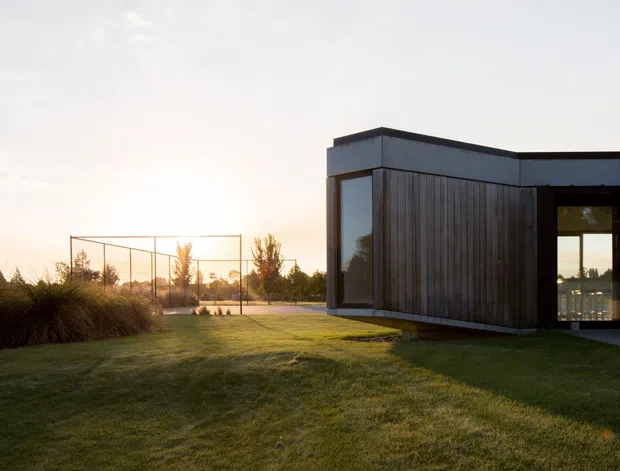 A low, minimalist modern building is photographed at dusk from a landscaped lawn. The dark, box-like structure has vertical timber cladding, dark metal or painted trim, expansive glazing, and a flat roof. A fenced recreational court is visible in the grounds, with warm sunset light and open lawn surrounding the building.