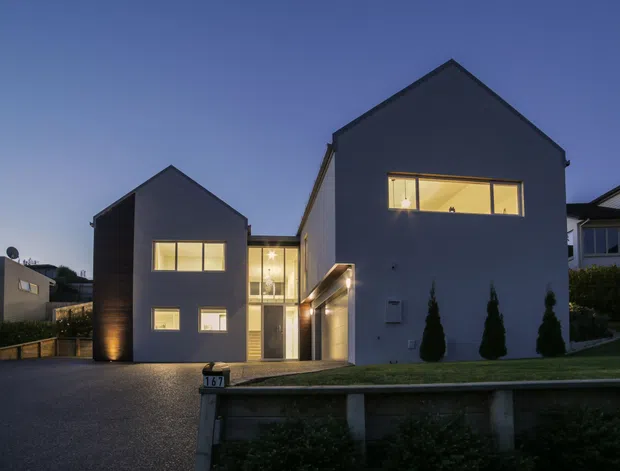 Twilight exterior view of a contemporary two-volume residential house with steep gabled roofs and dark grey rendered façades. Warm light glows from the windows and glazed entry connection, while a paved driveway, low retaining wall, lawn, and clipped conifer planting frame the front elevation.