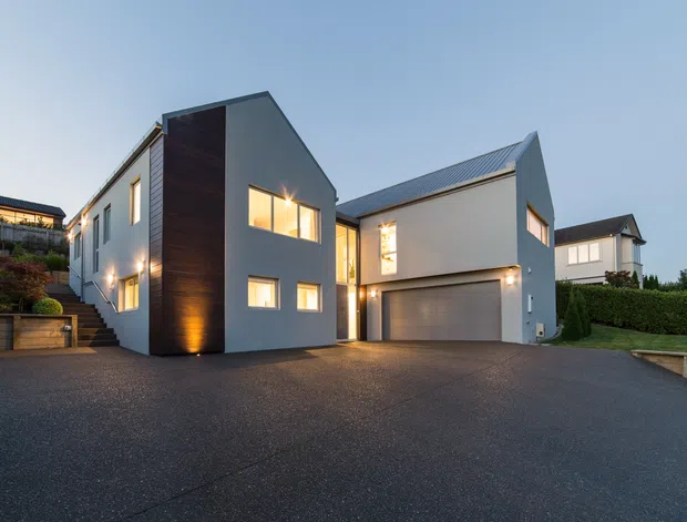 A contemporary multi-level residential home viewed from the driveway at dusk. The house has crisp white rendered walls, dark vertical timber cladding on a prominent entry volume, pitched metal roofs, large windows, and an integrated double garage. Landscaped terraced planting and neighbouring homes are visible around the paved driveway.